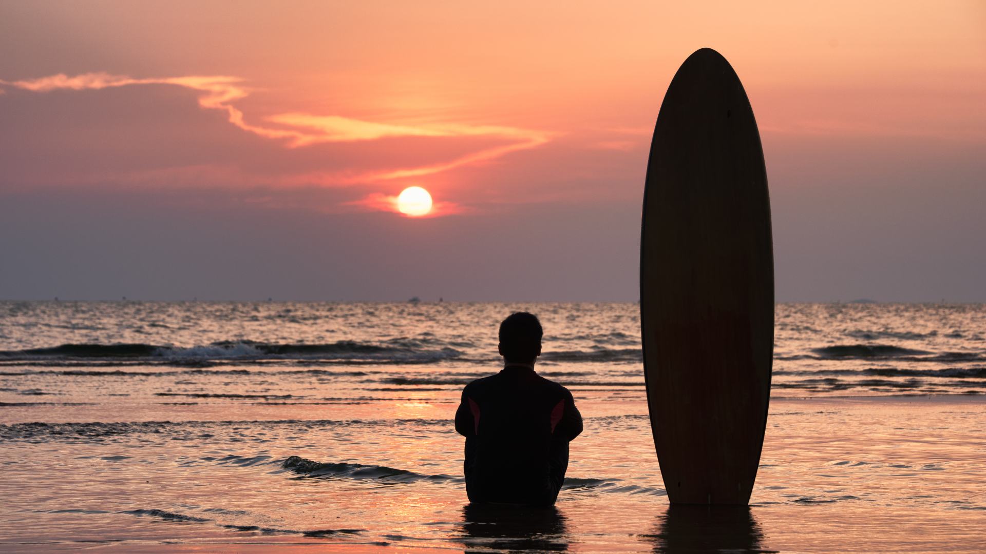 Ragazzo seduto di fronte al mare che ammira un tramonto con una tavola da surf di fianco